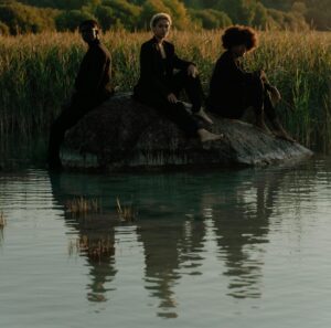 Three adults in black suits sitting on a rock, reflecting by a lake in nature.